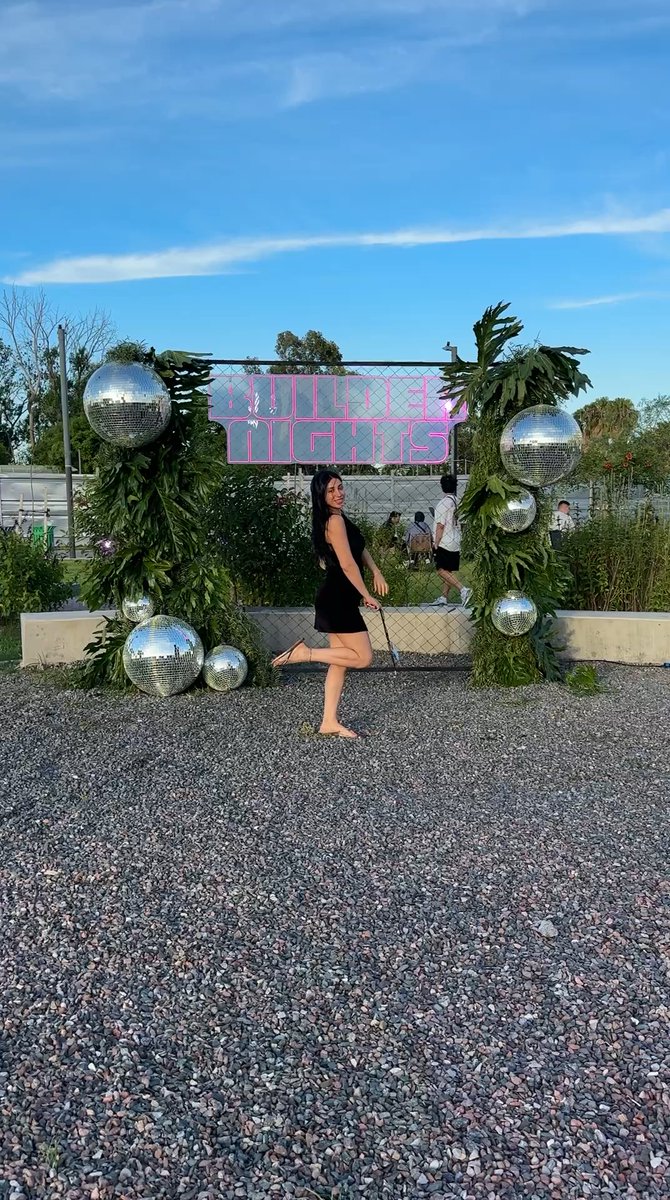 A woman in a black dress poses outdoors in front of a neon sign reading 'BUDDY LIGHTS' framed by greenery and disco balls.