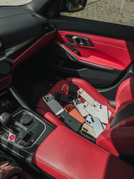 The interior of a luxury car with striking red leather seats and carbon fiber trim, with approximately nine smartphones in various colors piled on the passenger seat.