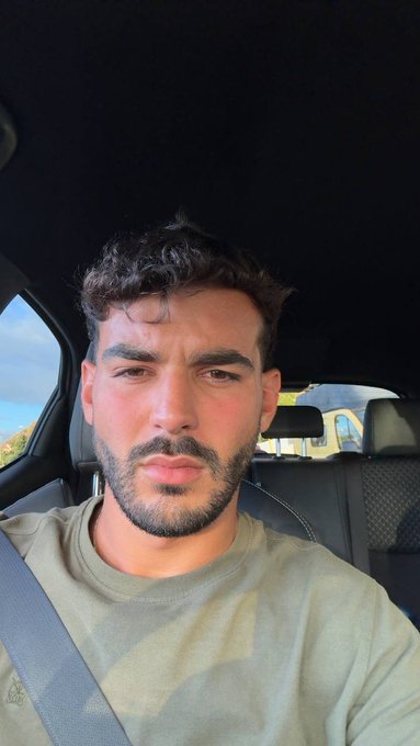 A selfie of a young man with curly dark hair and a beard, wearing an olive green t-shirt and seatbelt inside a car.