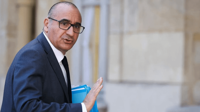A man in a dark suit and glasses, carrying a blue folder, glances back while walking through a grand marble building, likely a French government or legal institution—fitting imagery for the post about France's crackdown on crypto-related crimes and the arrest of an insider leaking investors' data.