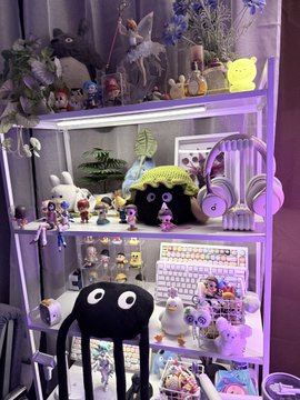 An illuminated display shelf filled with various anime figurines, plush toys, and collectibles including a crocheted Soot Sprite and Hello Kitty, lit with purple and white lights.