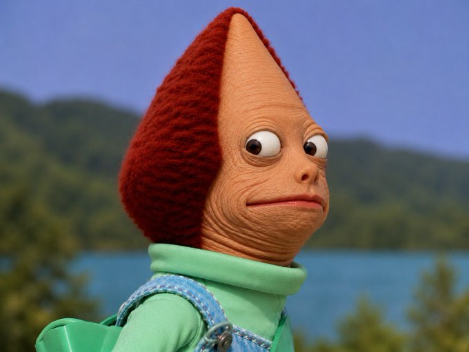 A close-up of a Patamon-like creature with a red fuzzy head, peach-colored features, and a green and blue outfit, set against a blurred background of trees and water.