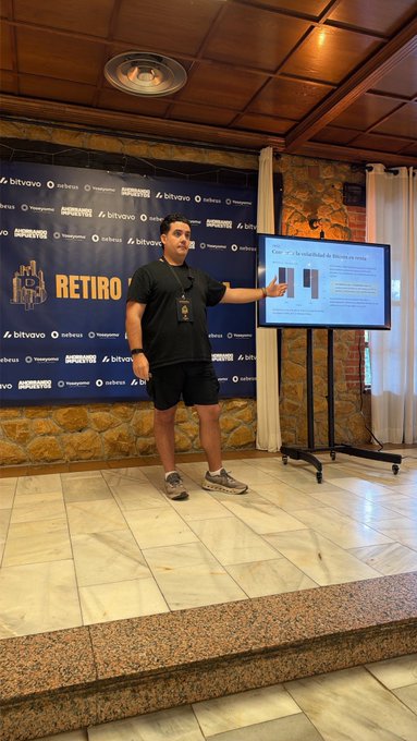 A presenter stands on stage at the 3rd Bitconia Retreat, pointing at a screen displaying a slide about bitcoin exposure options, with event branding and sponsor logos like "bitvalo" and "Nebula" visible on the backdrop behind him.