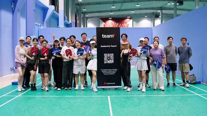 A large group of people pose together at a badminton event, holding paddles and standing behind a sign reading 'team1 Avalanche Team1' with a QR code in an indoor sports facility.