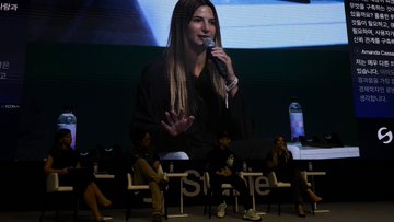 A panel discussion takes place on a stage with a large screen displaying a woman speaking into a microphone in the background. The hashtag "#SheTalks" is partially visible on the stage front.