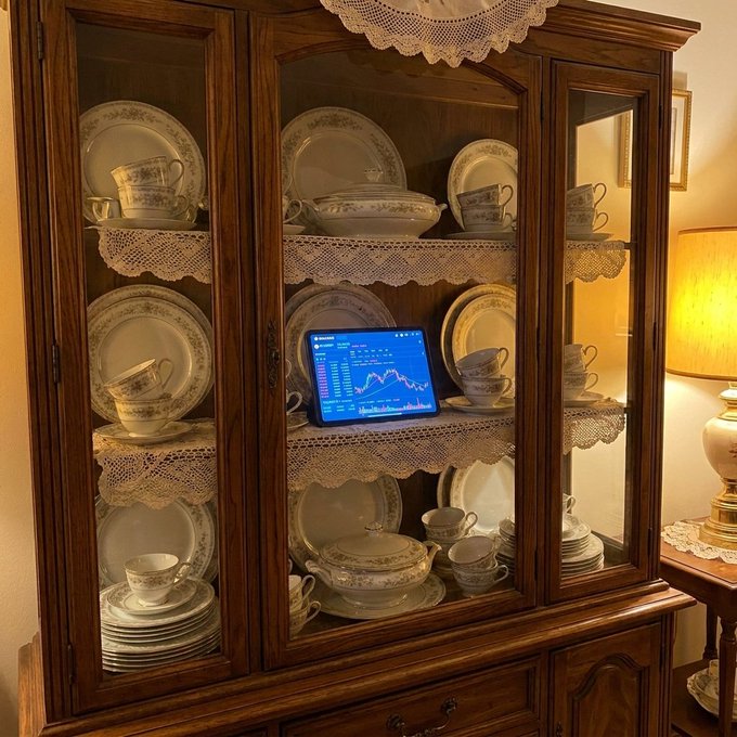 A wooden china cabinet displays porcelain dishware on lace doilies, with a tablet showing a blue stock/crypto chart placed incongruously among the plates.