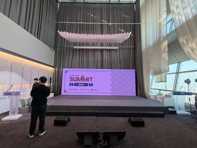 Event space setup for the 'SOLive SUMMIT Korea' with a large purple LED screen on a stage, decorative white ceiling installation, and a person standing in the foreground.