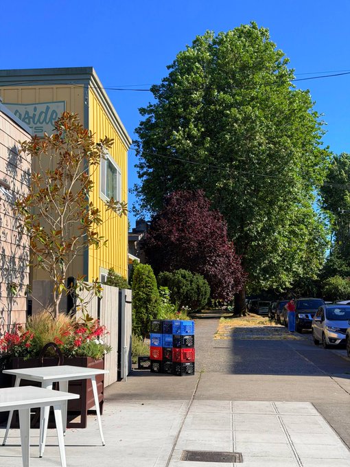 A sunny street scene featuring a yellow building with a 'Slide' sign, outdoor seating with white tables, magnolia trees, and stacked blue and red crates on the sidewalk.