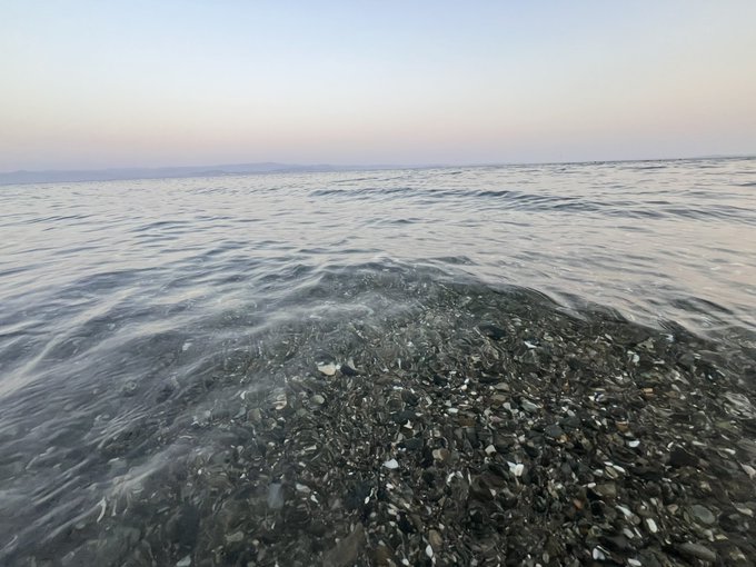 Low-angle photograph of calm sea water gently washing over a pebbled shoreline, with soft pastel sky tones of dusk or dawn visible in the background.