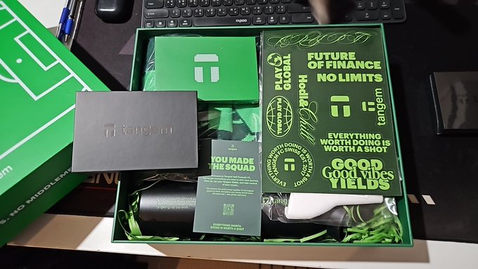 A green-themed promotional merchandise box containing cards with slogans like 'FUTURE OF FINANCE NO LIMITS', 'GOOD VIBES', and 'EVERYTHING WORTH DOING IS WORTH A SHOT', along with logos for Play Global and Tangem.