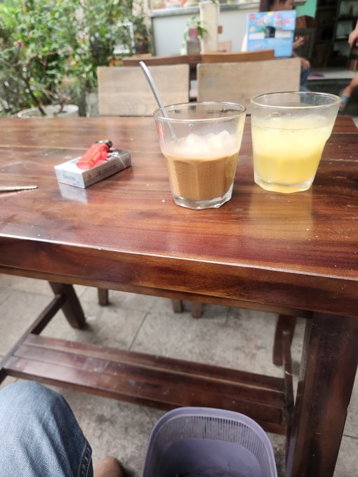 The image shows a wooden table with two glasses of drinks, one containing coffee and the other lemonade. There are also some items on the table such as a lighter, a cigarette, and a cup. The scene takes place outdoors at what appears to be a cafe or restaurant setting.