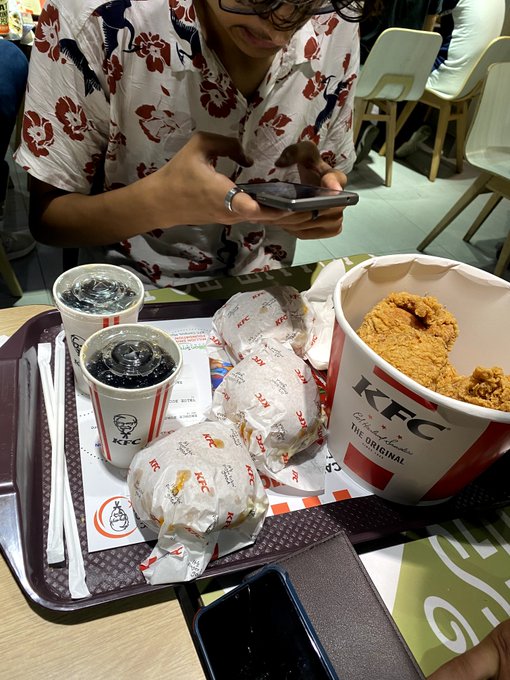 A person in a red and white floral shirt using a smartphone while seated at a KFC tray with fried chicken pieces, wraps, and two drinks.