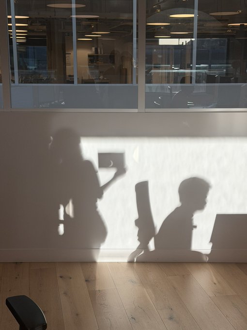 An artistic photograph showing the shadows of two people—one standing while holding up a phone, and another seated at a laptop—cast onto a white wall beside a large window.