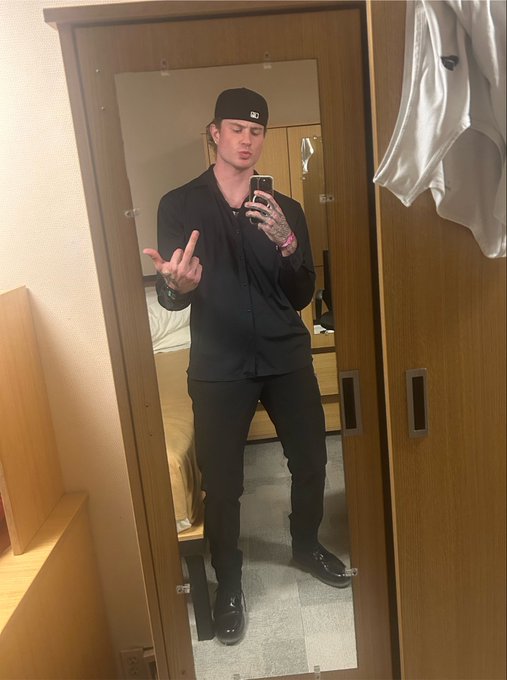 A mirror selfie of a man dressed entirely in black, wearing a cap, long-sleeve shirt, and pants, posing with a hand gesture in what appears to be a hotel room.