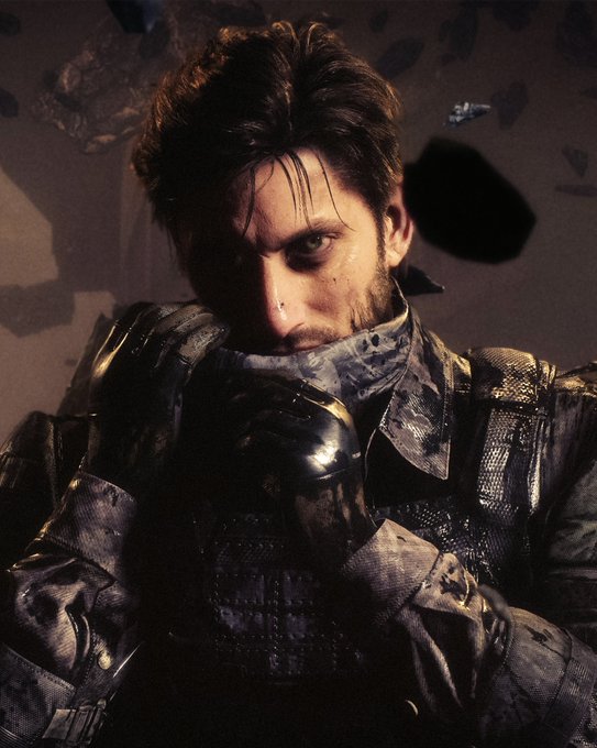 A man with tousled hair and a stubbled beard glares intensely forward, his gloved hands clutching a blood-stained mask up to his mouth against a dark, rocky backdrop. The image visually echoes the haunting tone of "Death Stranding 2," capturing the gritty, post-apocalyptic aesthetic central to Kojima Productions' narrative.