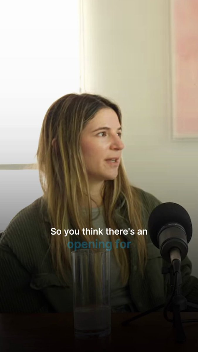 The image shows a woman sitting behind a microphone, likely participating in an interview or podcast recording session. She has blonde hair and is wearing a green jacket. There are two microphones visible in the scene - one being used by the woman and another placed on top of her head. The background features a white wall with a pink painting hanging above it.