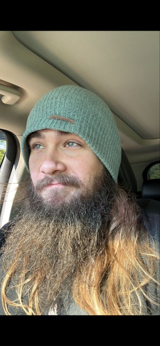 A man with a long brown beard wearing a green beanie sits inside a vehicle, looking off to the side.