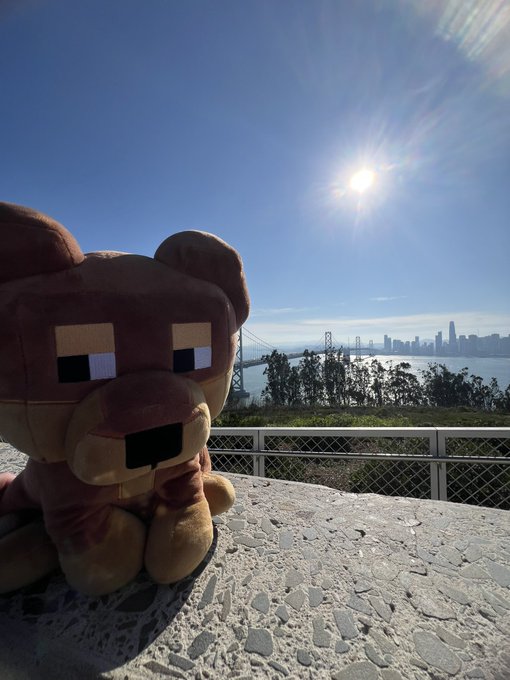 A pixelated plush bear sits on a stone ledge overlooking the San Francisco Bay Bridge and skyline, basking in bright morning sunlight that perfectly captures the "Gm" and "keep going" spirit of the post's message.