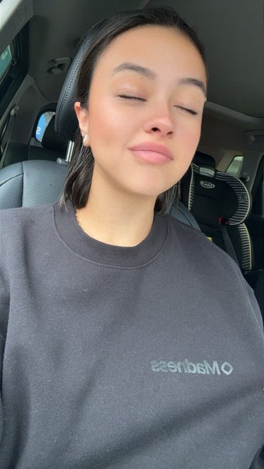 A selfie of a woman with dark hair and closed eyes wearing a dark gray sweatshirt with 'esrisbsMo' (mirror text) visible, sitting in a car with a child car seat visible in the background.