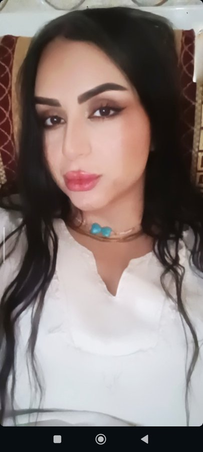 A woman with long dark hair, heavy eye makeup, pink lipstick, and a turquoise beaded necklace looks at the camera while wearing a white top.