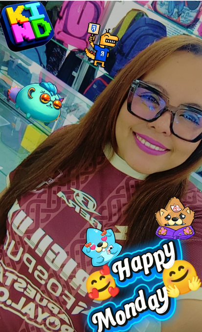 The image features a woman with long brown hair wearing glasses, smiling for the camera while posing for a photo. The background of the picture includes various cartoon characters and stickers related to the "Kiddie" brand. The caption of the image reads "Happy Monday and a wonderful start to June! Let this new month bring fresh energy, new opportunities, and endless reasons to smile @Kind_HQ @KanstarWorld @AngryDynomites @RoninInfinity @AxieInfinity #RoninMondaySelfie". The woman's glasses are prominently displayed in the image.