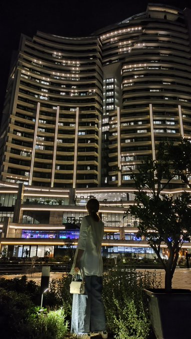 A nighttime photo of a person standing in a landscaped plaza in front of a large, modern, illuminated apartment complex with curved architecture and many lit windows.