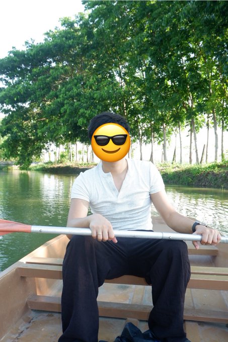 A person wearing a white shirt and dark pants sits in a boat holding a paddle, with their face covered by a sunglasses emoji. Trees and water are visible in the background.