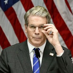 An older man in a suit and glasses adjusts his glasses in front of an American flag, appearing to be a government official.