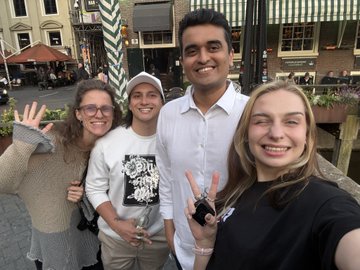 The image shows a group of four friends posing for a photo together outside an urban setting with buildings and trees in the background. The friends are standing close to each other, smiling and waving at the camera. One person in the group is holding a cell phone, possibly capturing the moment or checking something on their device. The overall atmosphere suggests that they have had a good time attending an event together and enjoying each other's company.