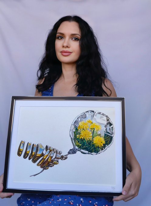 A woman with long dark wavy hair wearing a blue top, holding a framed artwork depicting a vintage lightbulb-shaped glass vessel containing yellow dandelions and a segmented caterpillar.