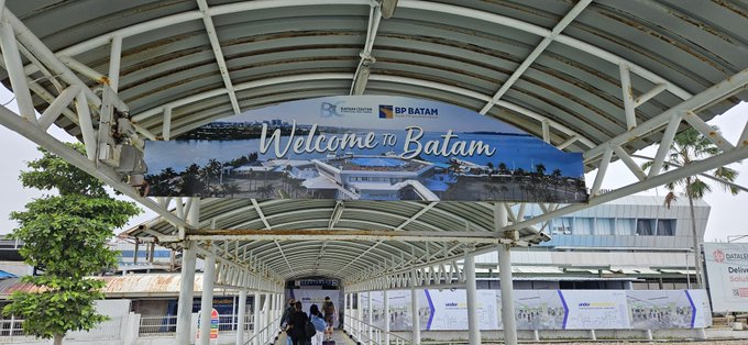 A covered walkway at a ferry terminal displays a 'Welcome to Batam' banner sponsored by BP Batam, with passengers walking toward the boarding area.