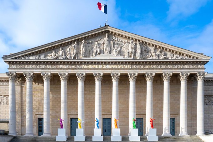 The facade of the Assemblée Nationale in Paris, featuring classical columns, a pediment, the French flag, and colorful modern sculptures lined up in front.