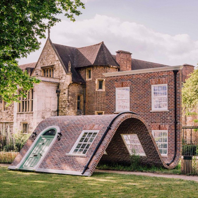 An arched brick building with a green door and windows sits on a lawn in front of a historic stone and brick manor house.