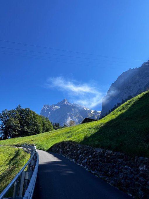 A scenic mountain landscape with a winding paved path, vibrant blue skies, green grassy slopes, and rocky peaks partially shrouded in clouds.