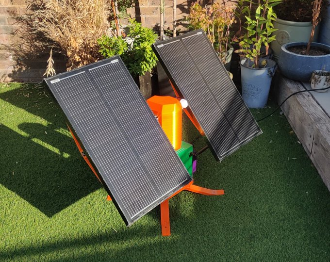 Two solar panels mounted on an orange stand are positioned on artificial grass in a garden setting, surrounded by potted plants.