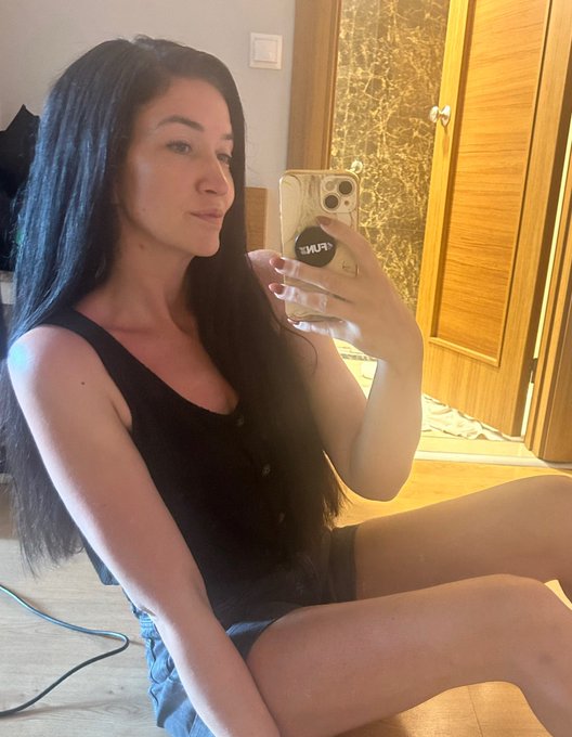 A woman with long dark hair takes a mirror selfie while holding a smartphone in a clear case that has the text 'WAIF' visible.