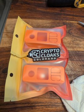 A sealed plastic package labeled 'CRYPTO CLOAKS COLORADO' containing two orange 3D-printed items, resting on a wooden surface.