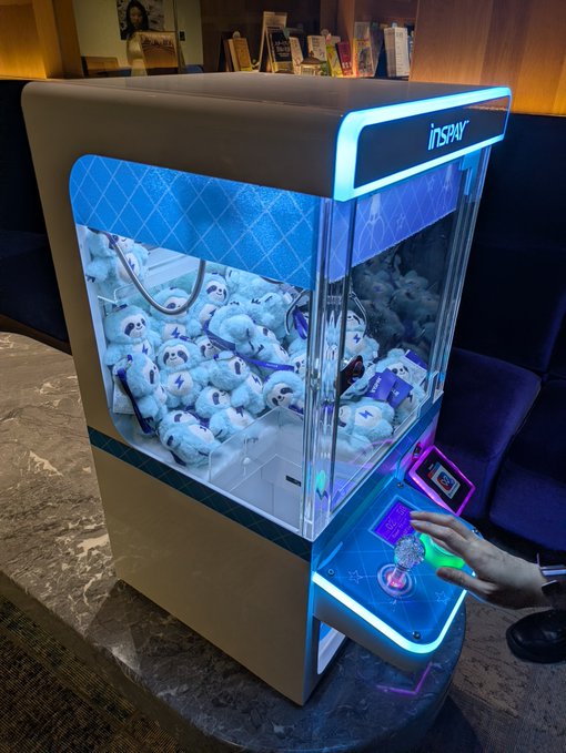 A glowing claw machine branded "INNSPAY" filled with plush toys accepts JPYC payments, with a hand inserting a coin into its illuminated slot, illustrating the innovative crypto payment system drawing enthusiastic attention.