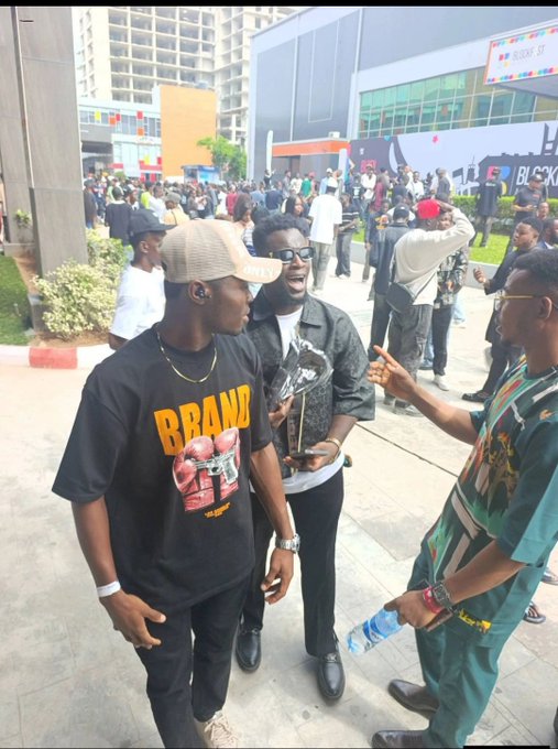 Three young men converse at an outdoor event, with one holding a trophy and wearing a black "BRAND" t-shirt, while "BLOCKFEST" signage is visible in the background, capturing the lively atmosphere of the African crypto/blockchain gathering.