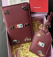 Burgundy leather wallets and luggage tags, embossed with colorful charm illustrations of vehicles, palm trees, and the Bitcoin "₿" symbol, are displayed alongside branded "Merch Mania" packaging, serving as a creative crypto-themed giveaway perfect for industry conferences.