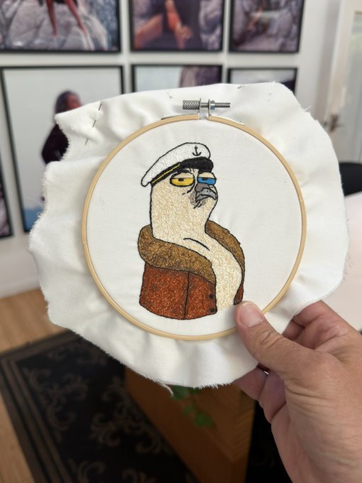 A hand holds up an embroidery hoop featuring a stitched portrait of a sailor/seagull character wearing a white cap and brown jacket, displayed in front of framed artwork.