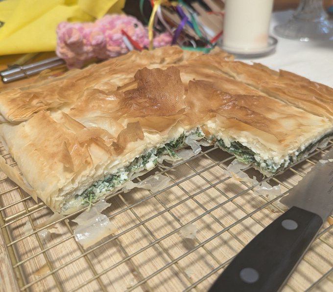 A golden-brown phyllo pastry pie filled with spinach and cheese sits on a wire cooling rack next to a knife.