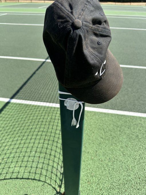 A black baseball cap sits atop a green tennis net post on a tennis court, with a white drawstring tied around the post.