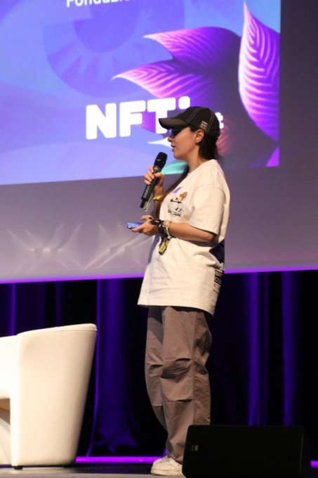 A person wearing a cap, sunglasses, and a white t-shirt speaking into a microphone on stage with a purple backdrop displaying the text 'NFT'.