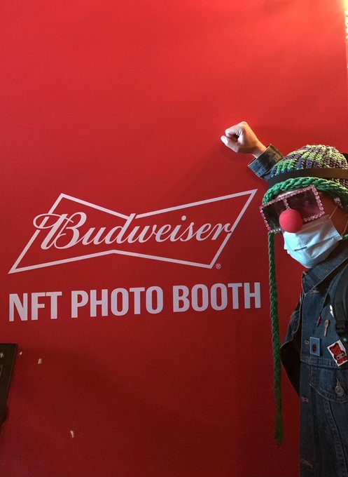 A person in a clown nose, mask, knit hat, and pink glasses poses with a raised fist in front of a red backdrop displaying the 'Budweiser NFT PHOTO BOOTH' logo.