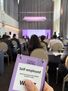 A hand holding a 'SUMMIT Blockchain Researcher & Web3 Certified Creator' badge labeled 'Self-employed' at a conference event.