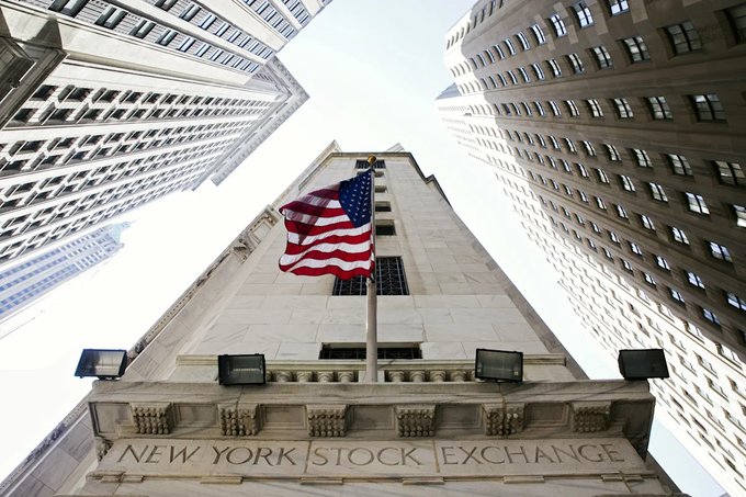 A dramatic low-angle shot looking up at the New York Stock Exchange building, featuring the American flag on a flagpole against a backdrop of tall skyscrapers and sky.