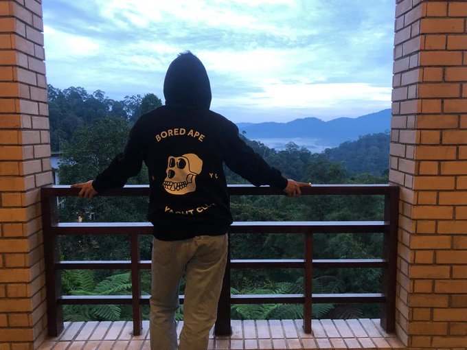A person wearing a black 'BORED APE YACHT CLUB' hoodie stands on a balcony overlooking a forested valley and distant mountains at dusk.