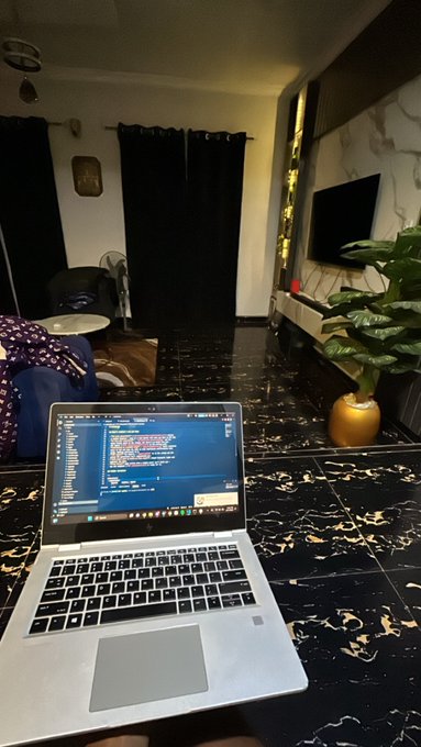A laptop open to a code editor displaying programming, sitting on a black marble-patterned floor in a dimly lit living room with a TV and potted plant in the background.