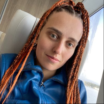 A selfie of a young woman with long reddish-orange braided hair wearing a blue jacket, taken inside a vehicle.